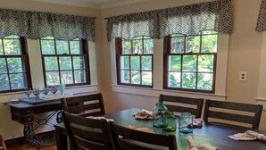 A dining area with a wooden table and chairs, a sewing machine, and windows with patterned valances.