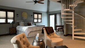 Une chambre à coucher comprenant un lit, un ventilateur de plafond, une fenêtre, une table de chevet, une lampe, une chaise, une petite table et un escalier.