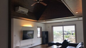 A spacious living room with a flat-screen TV, a ceiling fan, and a large sliding glass door leading to a garden.