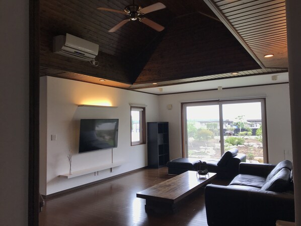 A spacious living room with a flat-screen TV, a ceiling fan, and a large sliding glass door leading to a garden.