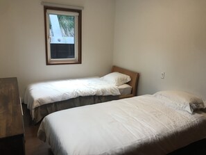 A small, clean room with two single beds, a wooden desk, and a window with a view of greenery.
