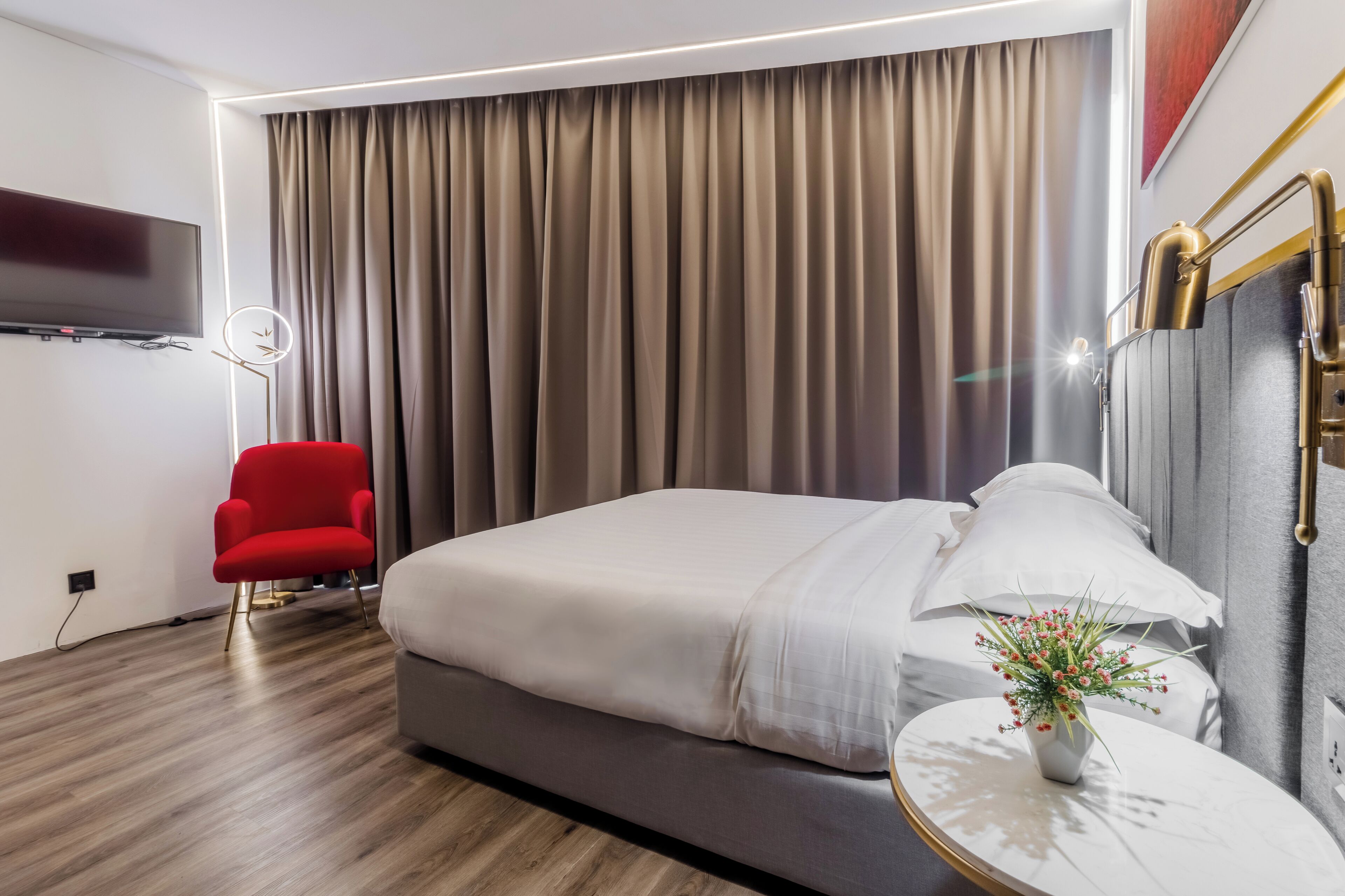 A modern hotel room with a large bed, a red armchair, a round table with a vase, and a flat-screen TV mounted on the wall.