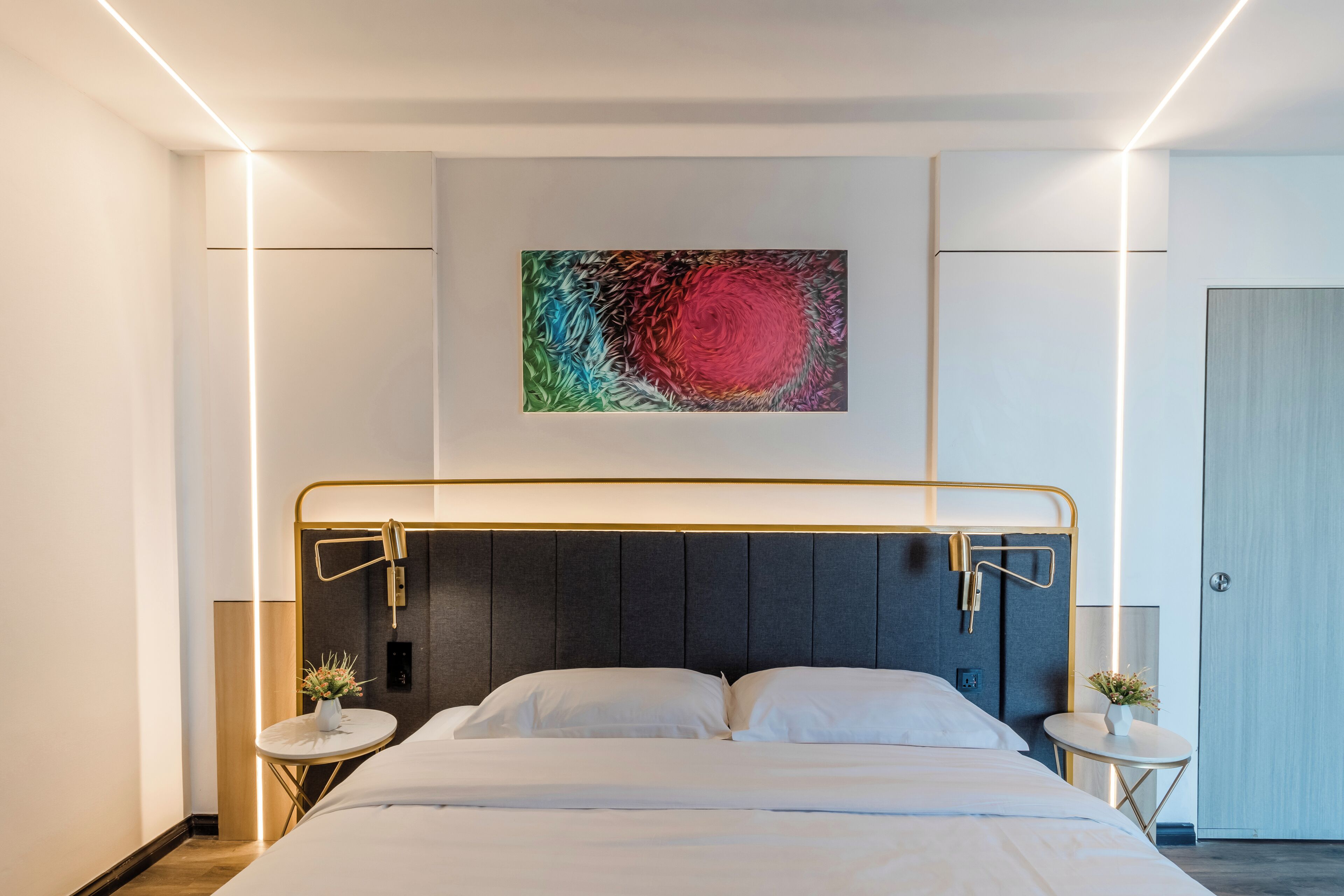 A modern hotel room with a bed, bedside tables, and a colorful abstract painting on the wall.