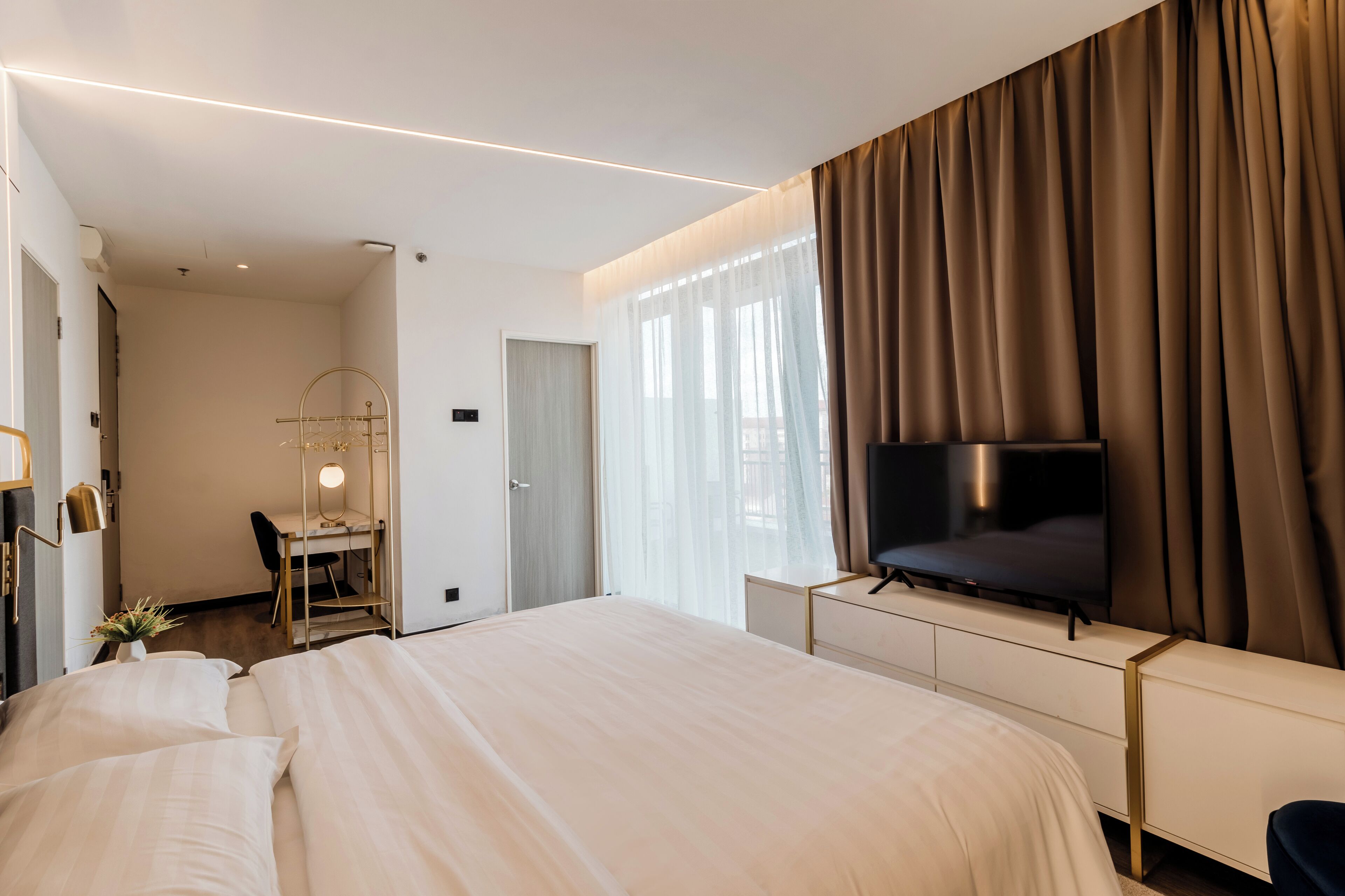 Una habitación de hotel moderna con una cama grande, un escritorio con espejo, televisión y un amplio ventanal con cortinas.