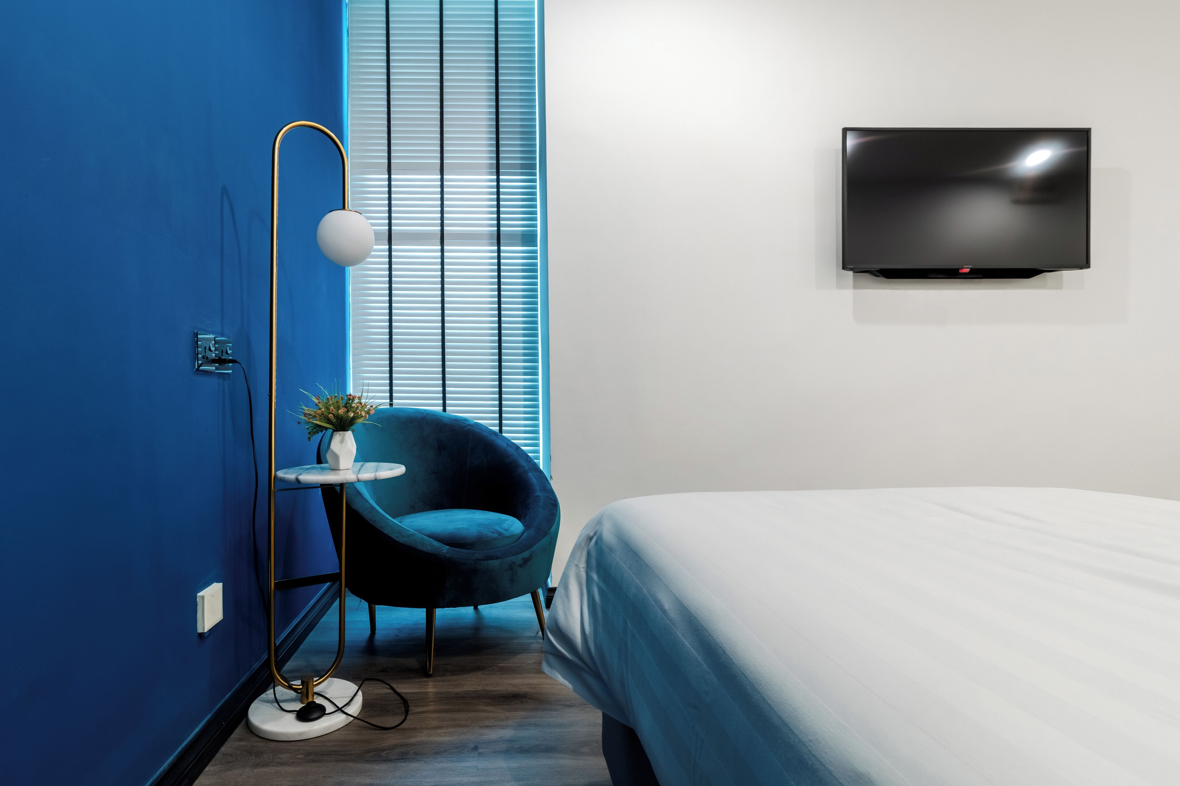 Une chambre d’hôtel moderne avec un mur d’accent bleu, un lit, un téléviseur fixé au mur, une lampe sur pied et une chaise avec une petite table.
