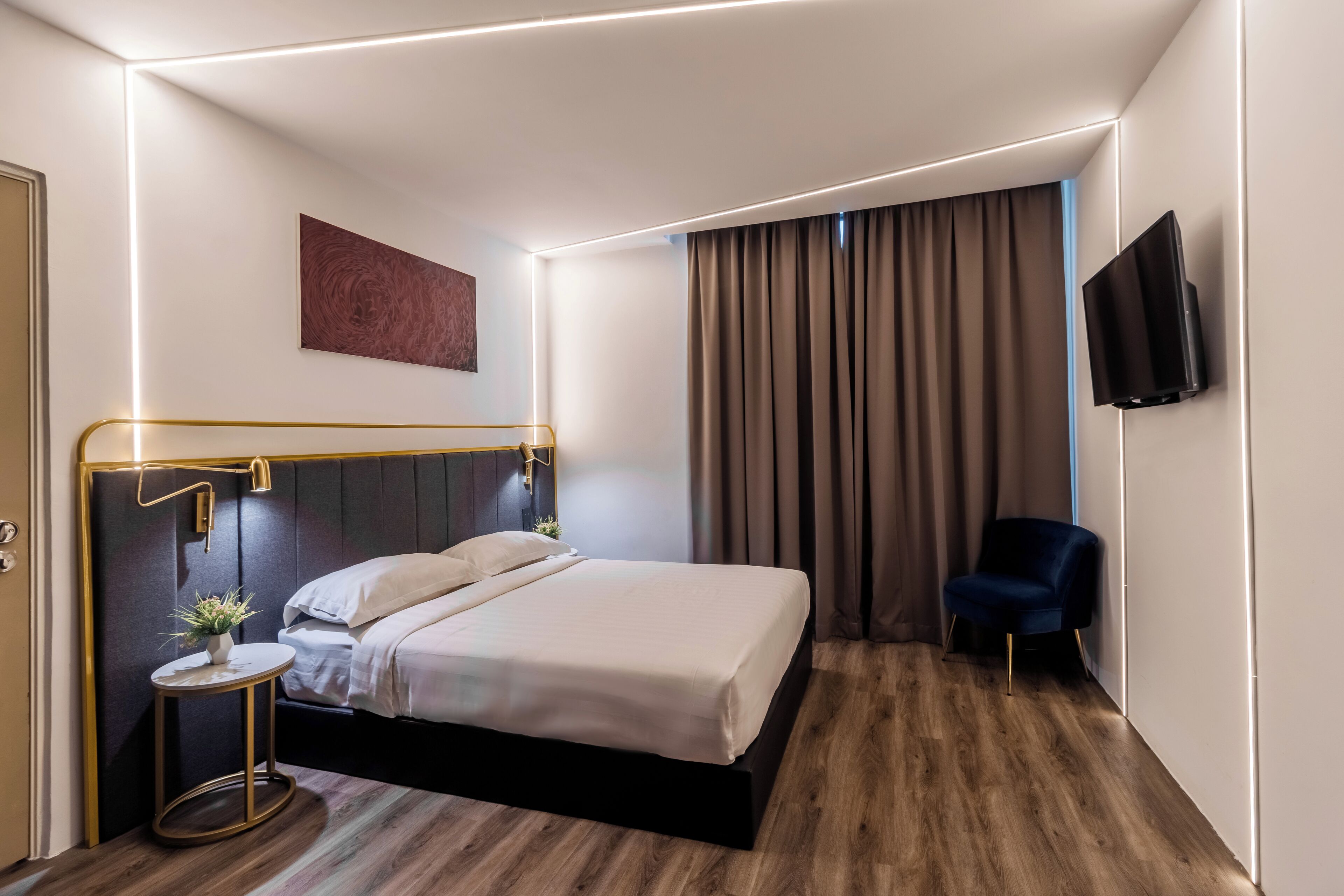 A modern hotel room with a neatly made bed, a small side table with a plant, a dark blue chair, and a flat-screen TV mounted on the wall.