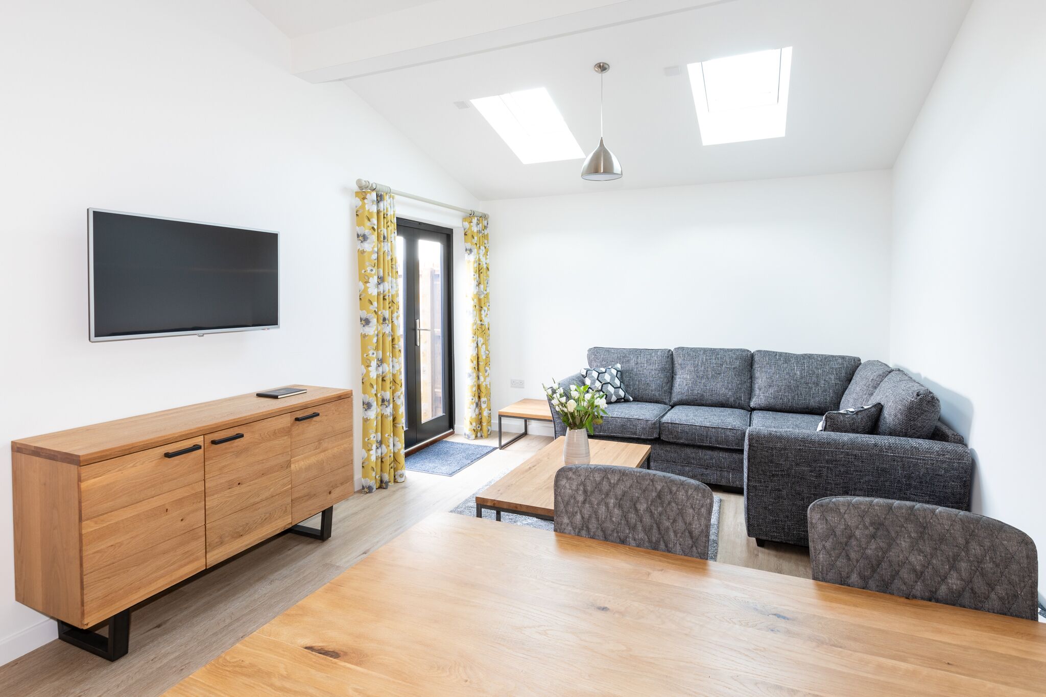 A modern living room with a wooden dining table, grey sofa, and a flat-screen TV mounted on the wall.