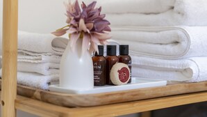 Un estante de madera con toallas blancas dobladas con cuidado, un florero blanco con una flor púrpura y tres botellas de productos para el cabello.