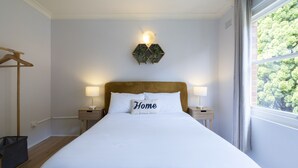 Una cama bien hecha con una almohada que dice 'Home', dos lámparas de noche, una mesita de madera y una planta colgada en la pared.