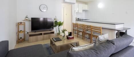 A modern living room with a sofa, a coffee table, and a TV. There is a kitchen area with a stove and cabinets.