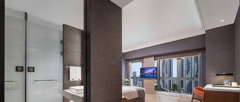 A modern hotel room with a large bed, a desk, and a view of the cityscape through large windows.