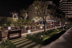 Un espacio al aire libre con mesas y sillas, rodeado de follaje e iluminado con luces colgantes.