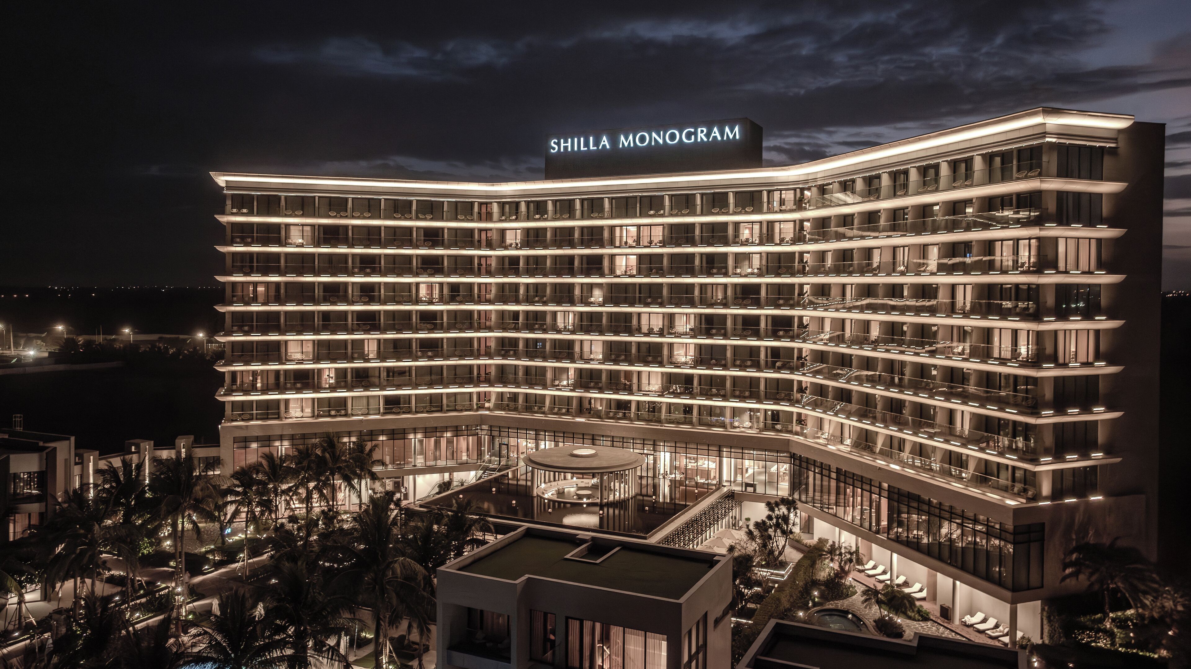 A multi-story hotel with a curved facade, named "Shilla Monogram." It features a rooftop pool area with lounge chairs and palm trees.