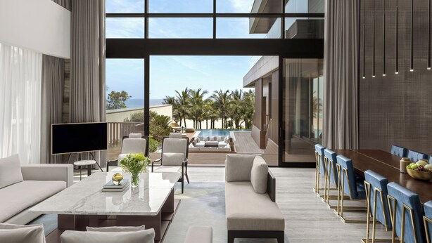 A modern living room with a large TV, comfortable seating, a dining area with blue chairs, and a view of a pool and palm trees. - Shilla Monogram Danang (Da Nang)
