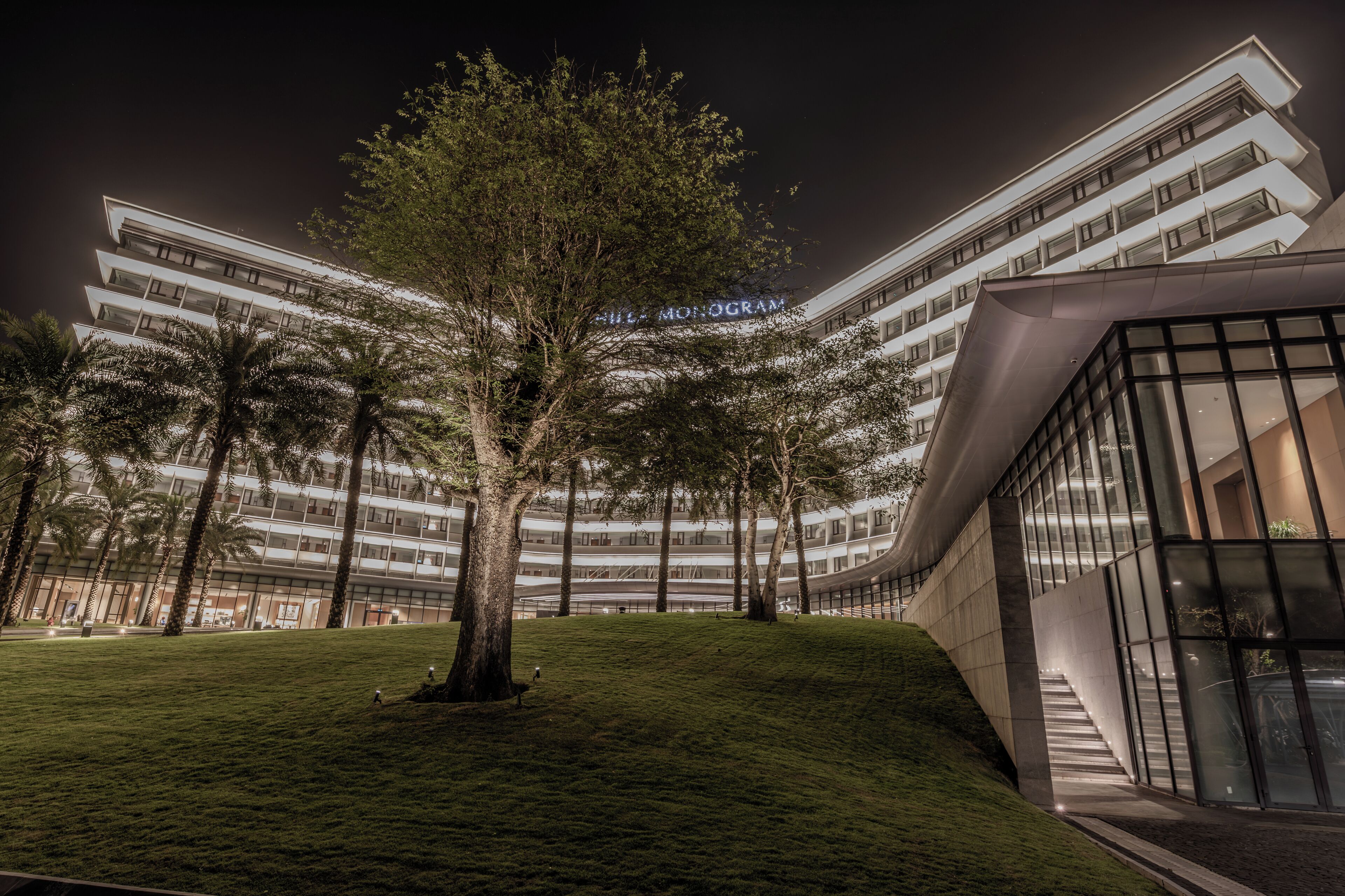 A modern hotel with a curved facade, a grassy area with trees, and a visible sign reading "Hotel Monogram."