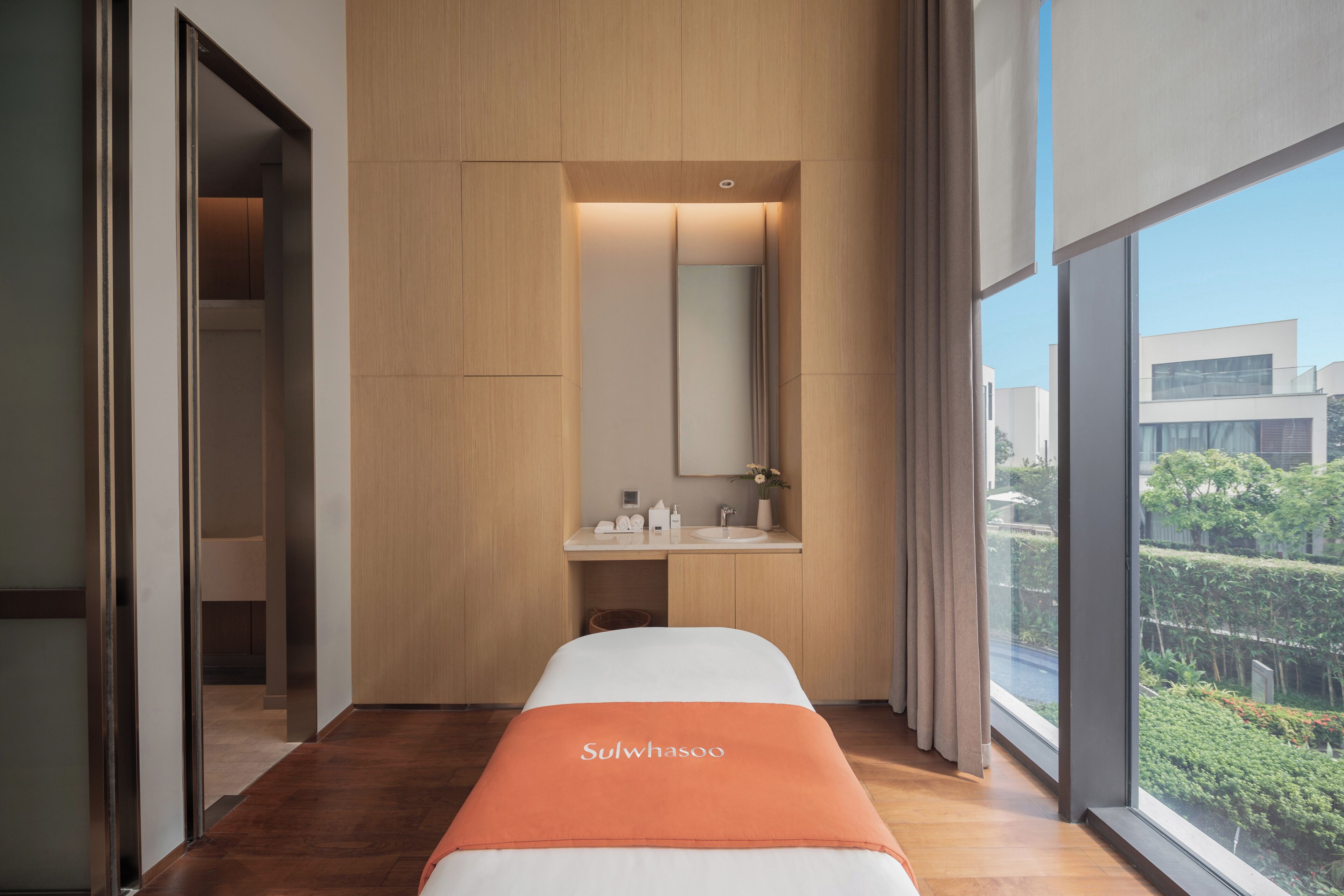 A modern hotel room with a bed, a bathroom with a sink and mirror, and a large window offering a view of the city.