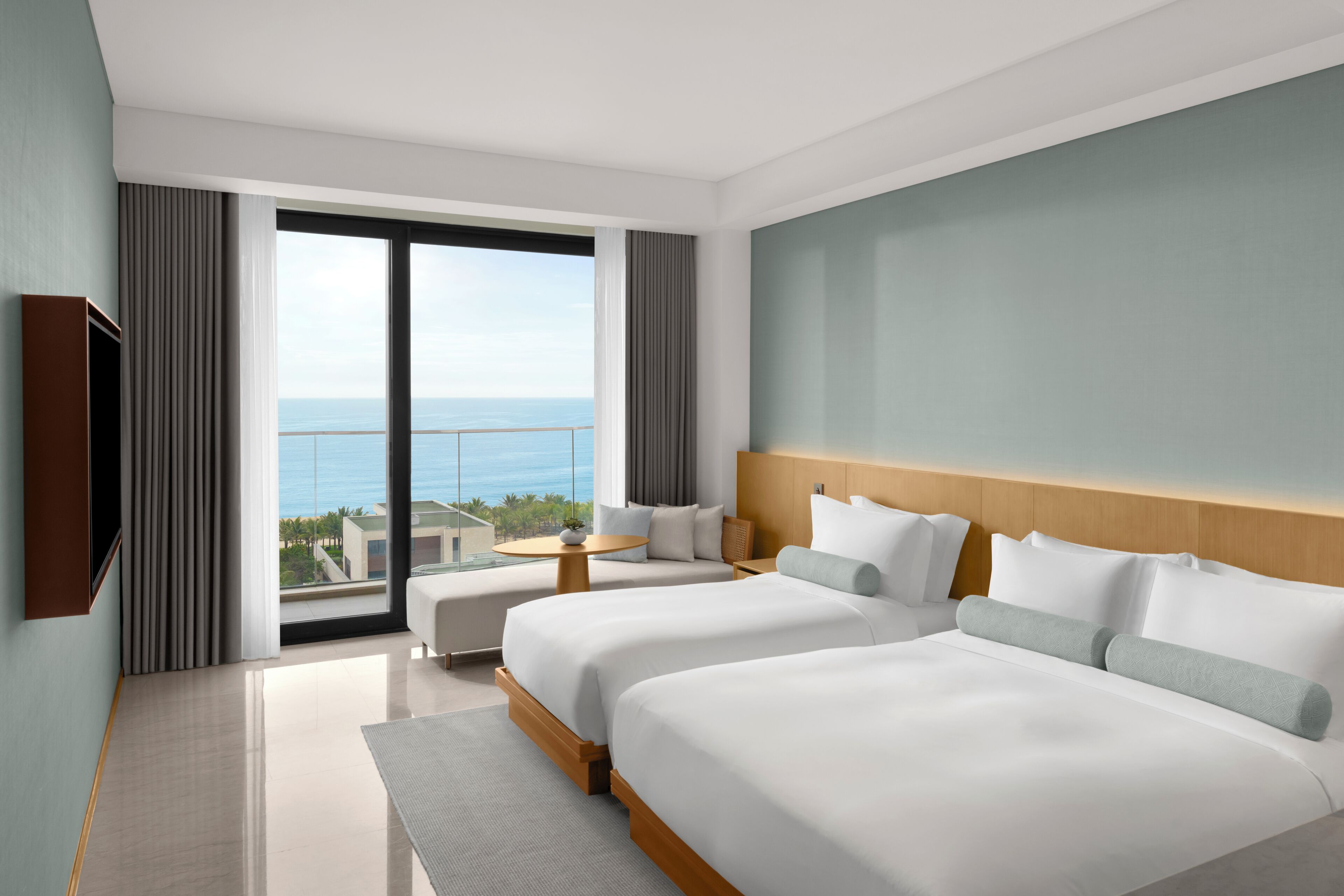 A hotel room with two beds, a TV, a balcony with a view of the sea, and a small table.