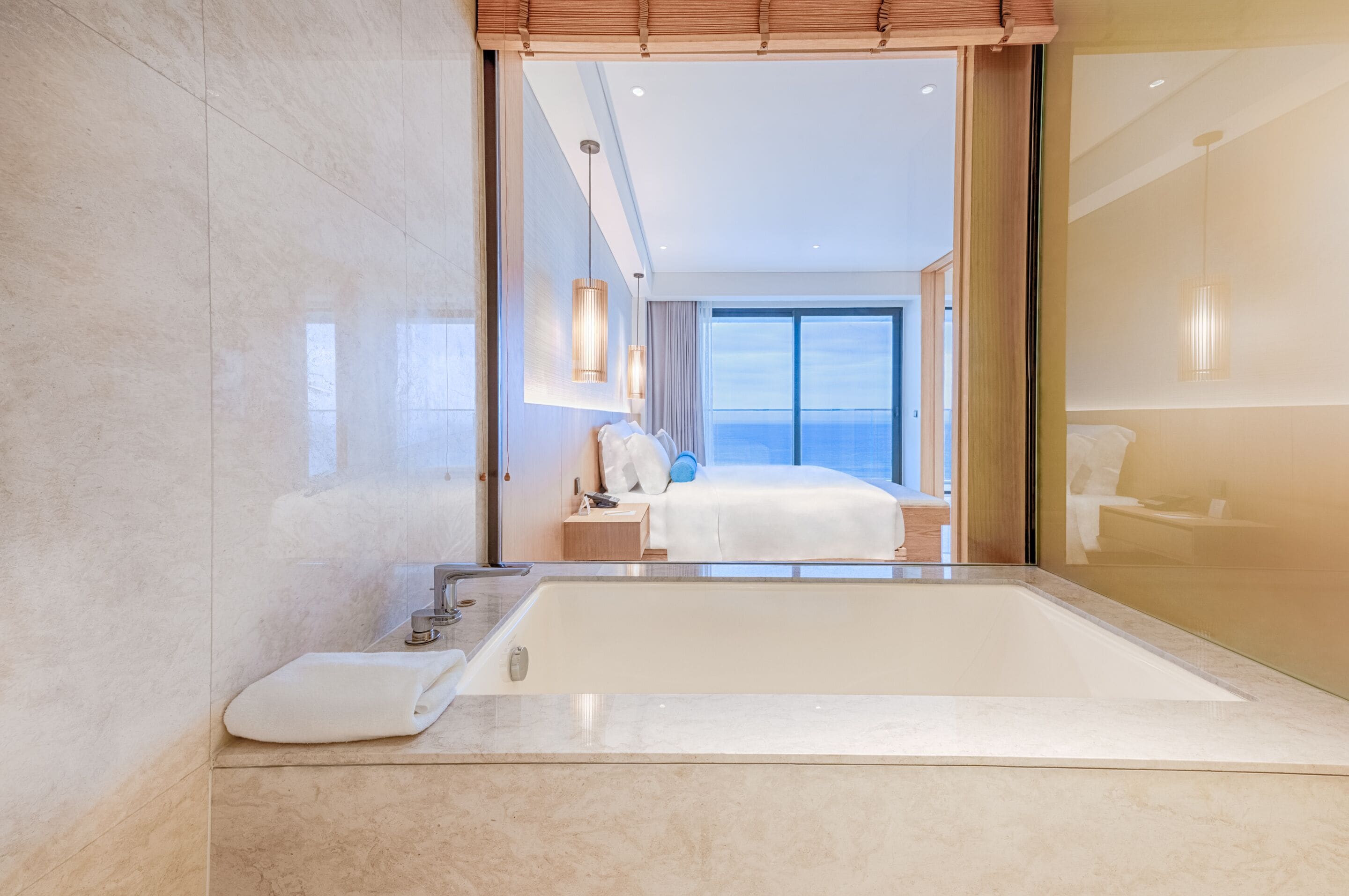 Un baño moderno con una bañera grande, un espejo y vistas a un dormitorio con cama y mesita de noche.