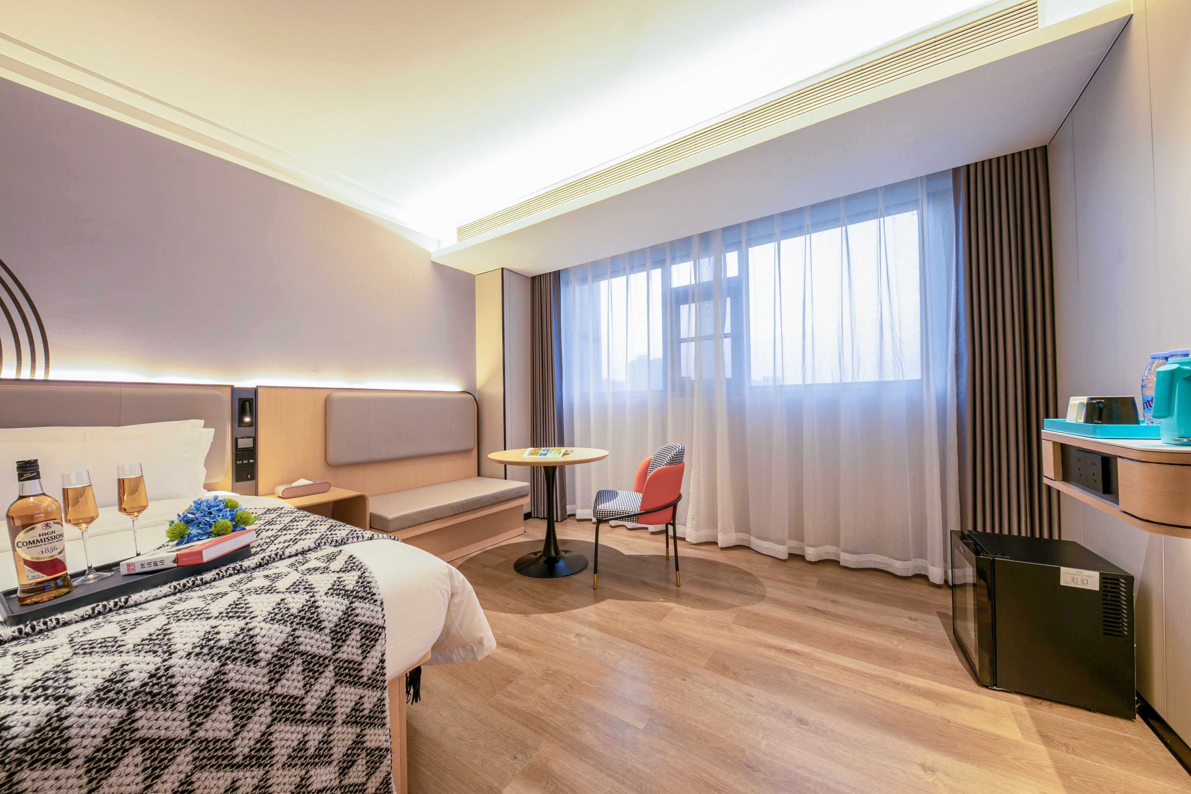 A hotel room with a bed, a small table, a chair, a sofa, a window with curtains, and a minibar.