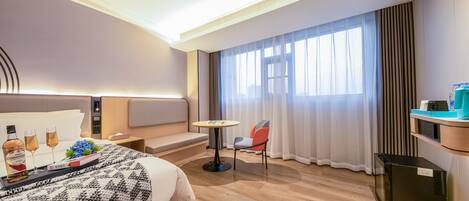 A hotel room with a bed, a small table, a chair, a sofa, a window with curtains, and a minibar.