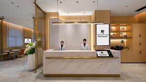 Zona de recepción de un hotel con mostrador de mármol, dos recepcionistas, un mostrador y un área de estar con una planta.