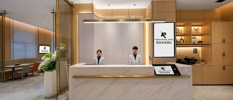 A hotel reception area with a marble counter, two receptionists, a reception desk, and a seating area with a plant.