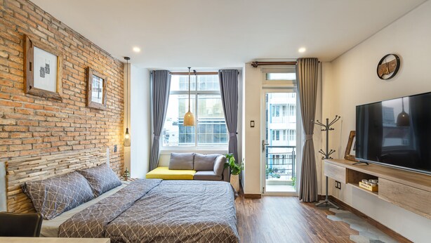 Un dormitorio moderno con pared de ladrillo, cama grande, televisor y balcón con cortinas.