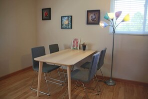 Une pièce avec un plancher en bois, une table de couleur claire et quatre chaises. Trois tableaux encadrés sont accrochés au mur.