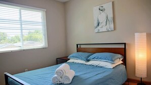Una cama bien hecha con ropa de cama azul y toallas blancas, una mesita de noche, un cuadro colgado en la pared y una lámpara de pie.