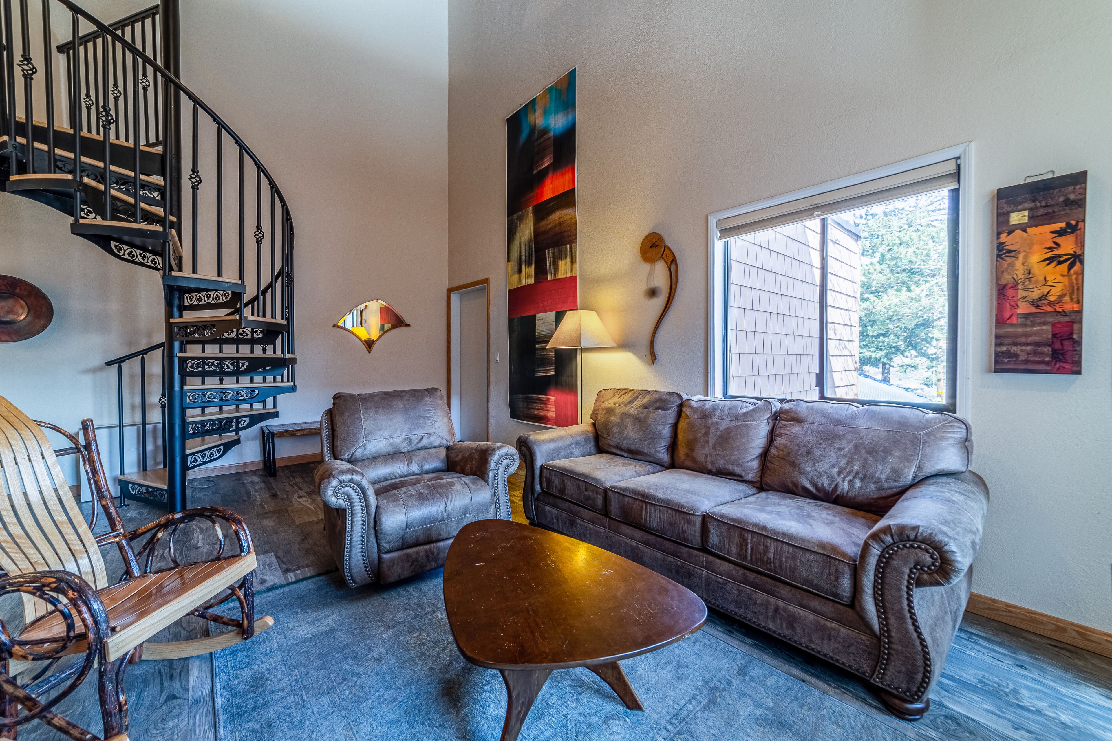 A living room with a brown leather sofa, two armchairs, a wooden rocking chair, a coffee table, a spiral staircase, and a large abstract painting.