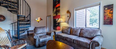 A living room with a brown leather sofa, two armchairs, a wooden rocking chair, a coffee table, a spiral staircase, and a large abstract painting.