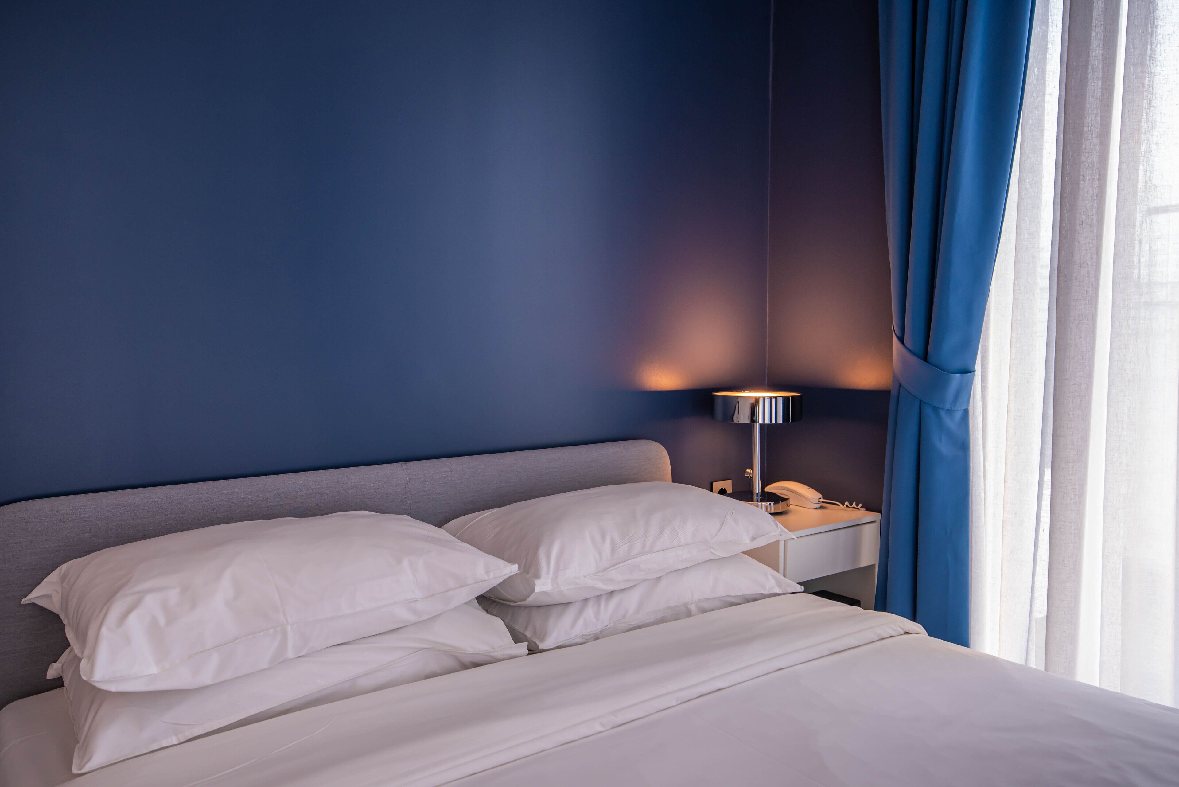 En pænt redt seng med hvide sengetøj i et rum med blå vægge og et natbord med en lampe.