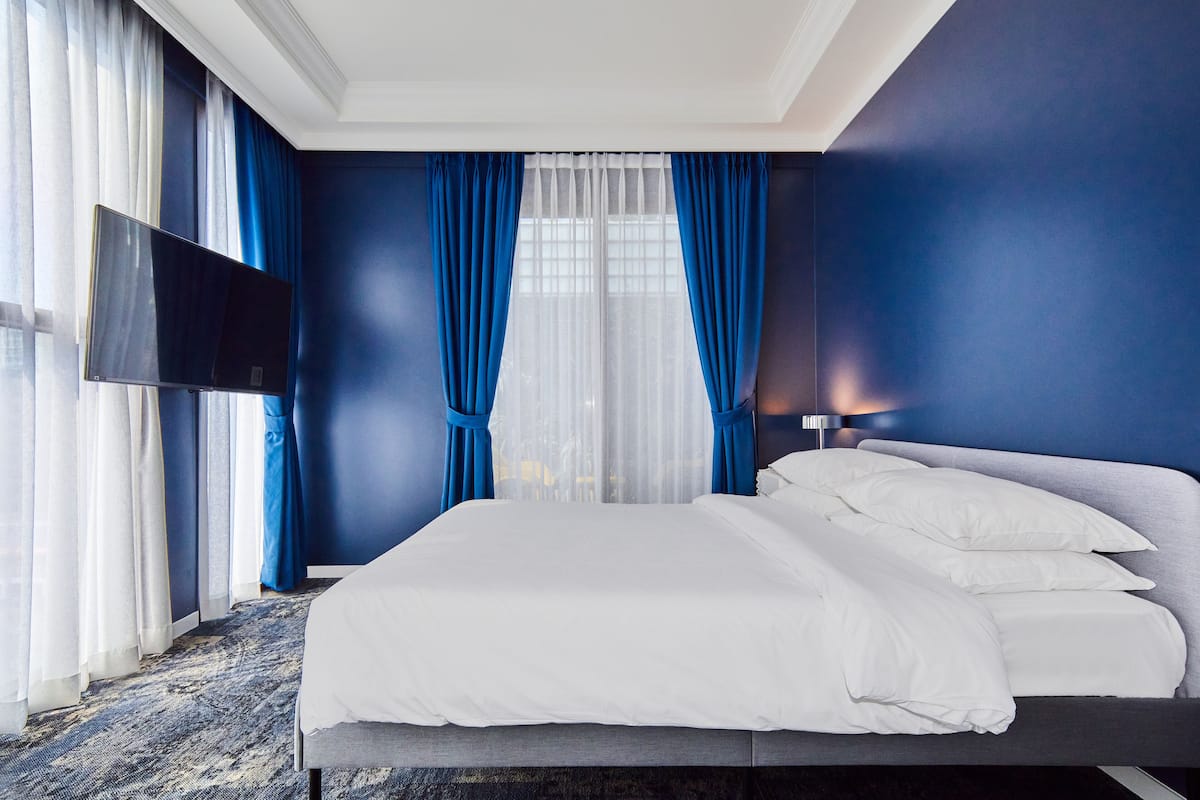 A modern hotel room with a large bed, a flat-screen TV, and a view of the outdoors through sheer curtains.