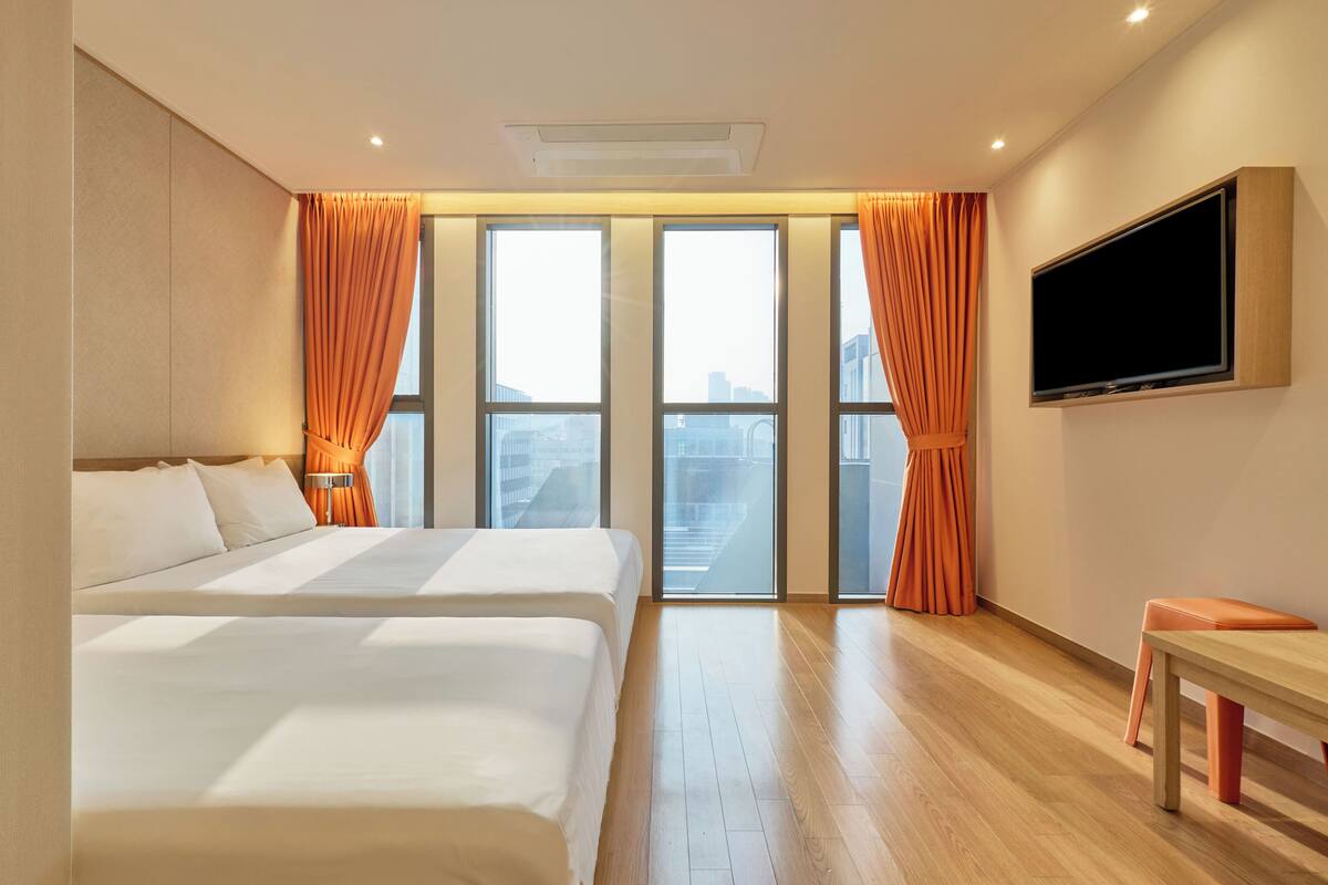 A hotel room with two beds, a TV, and large windows with curtains.