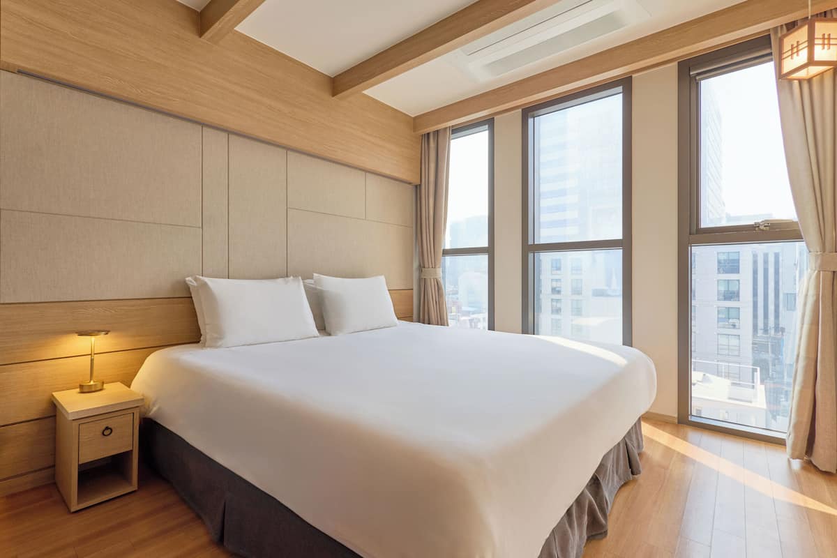 A hotel room with a large bed, a nightstand, a window with a view of buildings, and a wooden ceiling.