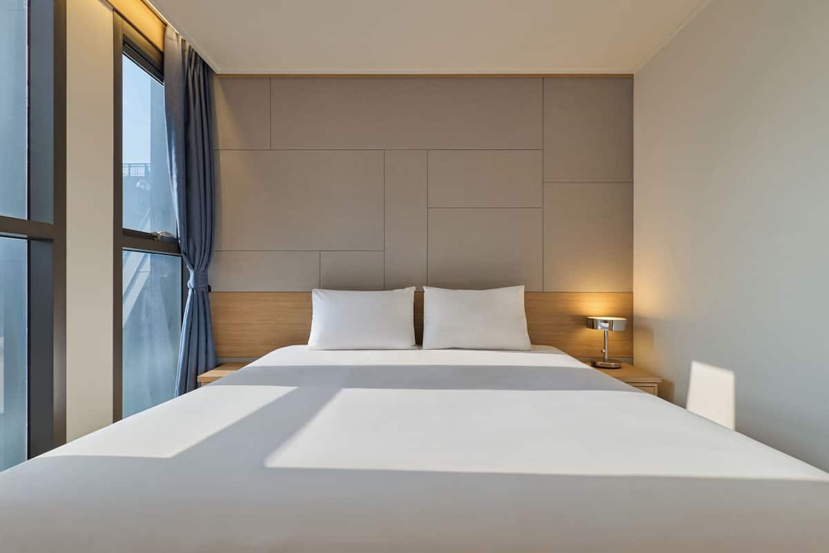 A modern hotel room with a large bed, a nightstand with a lamp, and a view through a window with curtains.