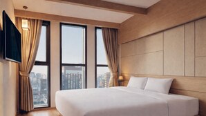 Una moderna camera d'albergo con un letto grande, vista sulla città attraverso finestre a tutta altezza, pannelli in legno sulle pareti e una TV a schermo piatto montata a parete.