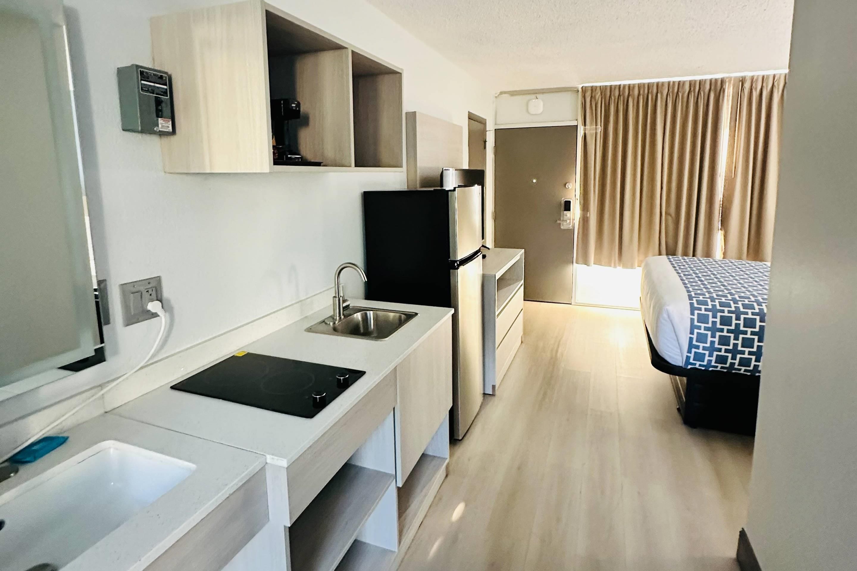 A compact hotel room with a kitchenette, a bed with patterned bedding, and a bathroom with a shower.