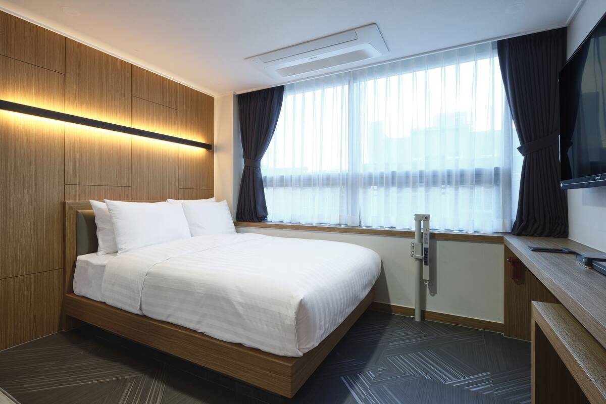 A modern hotel room with a large bed, wooden panel walls, a flat-screen TV, and a window with sheer curtains.