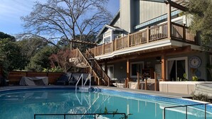 Een woonhuis met een zwembad, houten terras en een twee verdiepingen tellend huis met balkon.