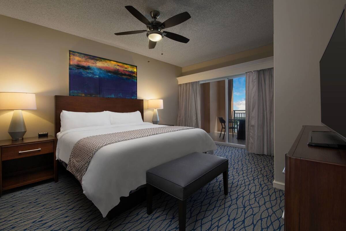 Ein Hotelzimmer mit einem Bett, zwei Nachttischlampen, einem Deckenventilator, einem großen Gemälde, einem Balkon mit einem Stuhl und einem Fernseher auf einer Holzkassette.