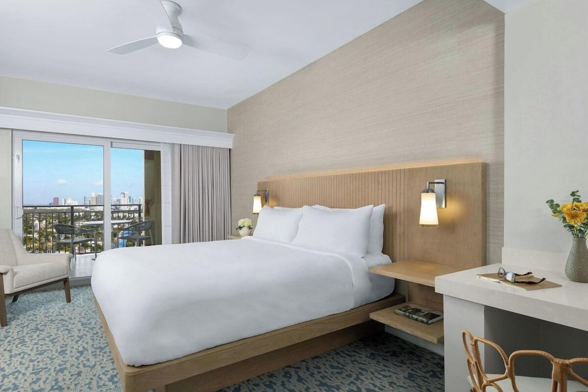 Una habitació d'hotel amb un llit, una taula de treball, una cadira, un balcó amb vistes i un ventilador de sostre.