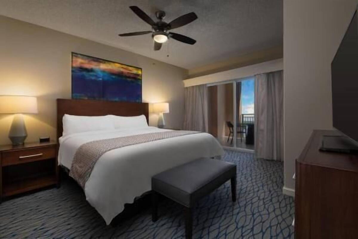 Ein Hotelzimmer mit Bett, Deckenventilator, Gemälde, Schreibtisch, Stuhl und Balkon.