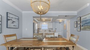 A dining area with a wooden table, chairs, and a chandelier. There is a kitchen in the background with a refrigerator and a window. - Top-Floor Gulf Views | Updated Condo Near Beach & Times Square & Pool Access (Fort Myers Beach)