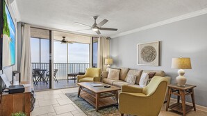 A spacious living area with a sofa, armchairs, a coffee table, and a side table with a lamp. A ceiling fan is visible, and there is a large window offering a view outside. - Top-Floor Gulf Views | Updated Condo Near Beach & Times Square & Pool Access (Fort Myers Beach)