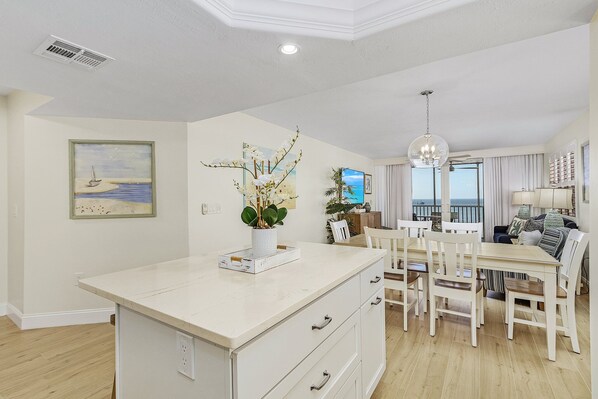A modern kitchen with white cabinetry, a central island, and a dining area with white chairs. - End Unit Beachfront Condo | Gulf Views & Walk to Times Square (Fort Myers Beach)