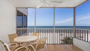 A balcony with a view of the beach, a round table with chairs, and a ceiling fan.