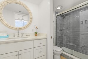 A bathroom with a white vanity, a round mirror, a small plant, and a glass-enclosed shower area. - End Unit Beachfront Condo | Gulf Views & Walk to Times Square (Fort Myers Beach)