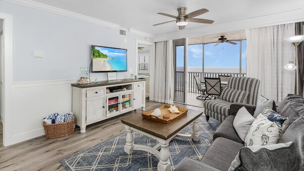 A spacious living room with a wooden coffee table, a striped armchair, and a sofa. A television is mounted on the wall, and there is a window with a view of the ocean. - Gulf-Front Condo | Heated Pool, Beach Access & Walk to Times Square (Fort Myers Beach)