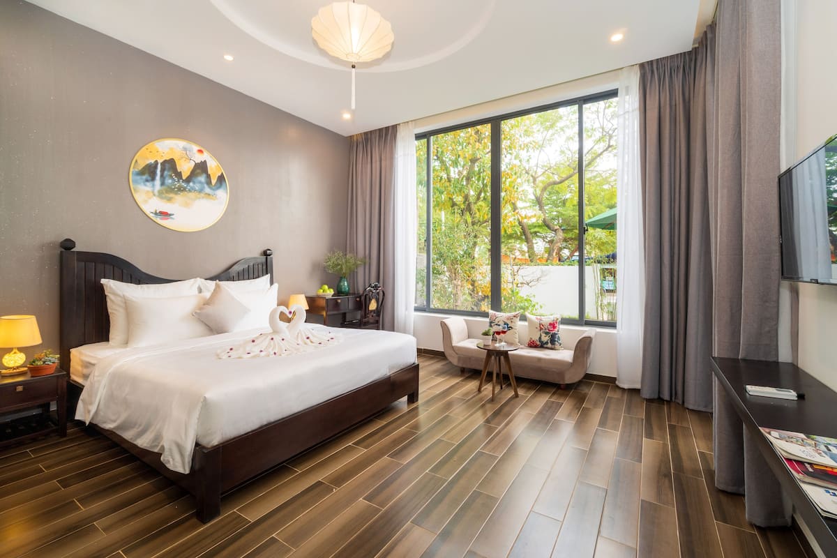 A modern hotel room with a large bed, a sofa, a TV, and a window with a view of trees.